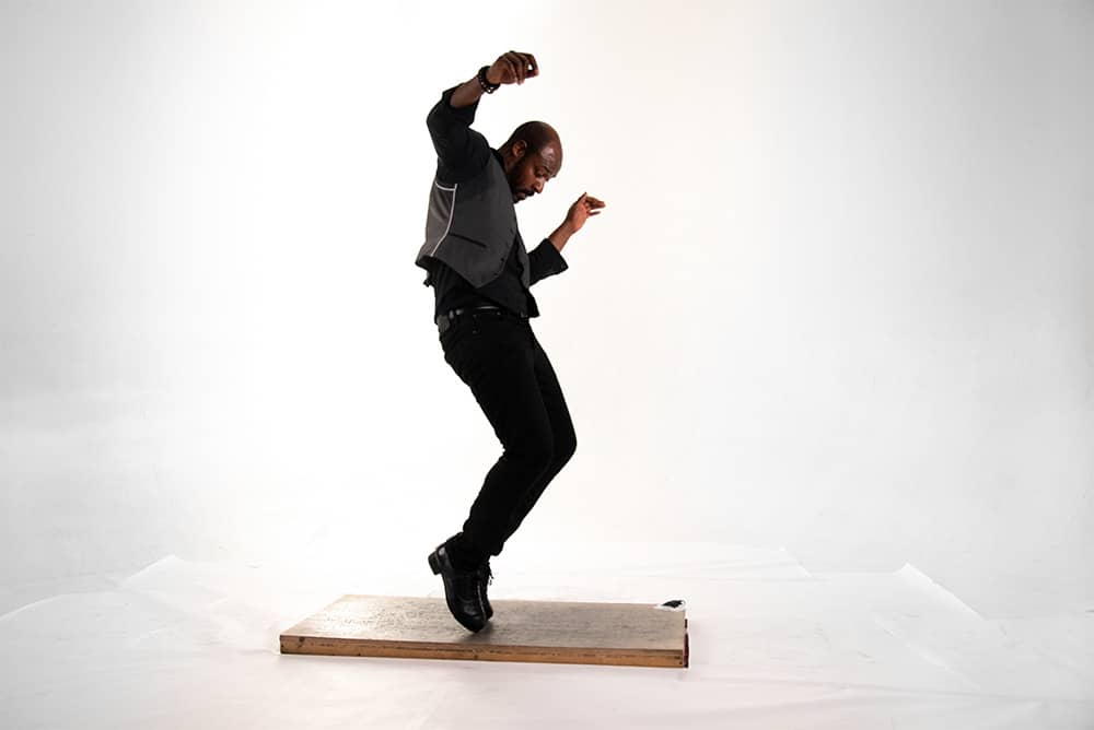 Description de l’image : 
Debout, une personne habillé tout en noir est sur la pointe des pieds. La personne touche une plateforme au sol et fait des mouvements de danse avec les bras en l’air et les genoux pliés.