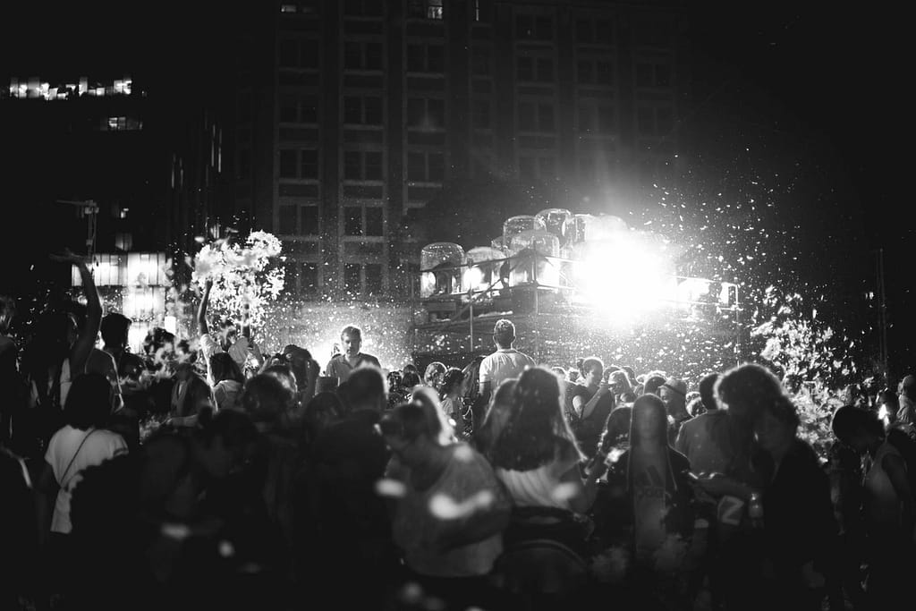 Description de l’image : L’image est une photographie en noir et blanc montrant une foule de personnes devant un spectacle en extérieur. En arrière plan, il y a les fenêtres d’un bâtiment. 