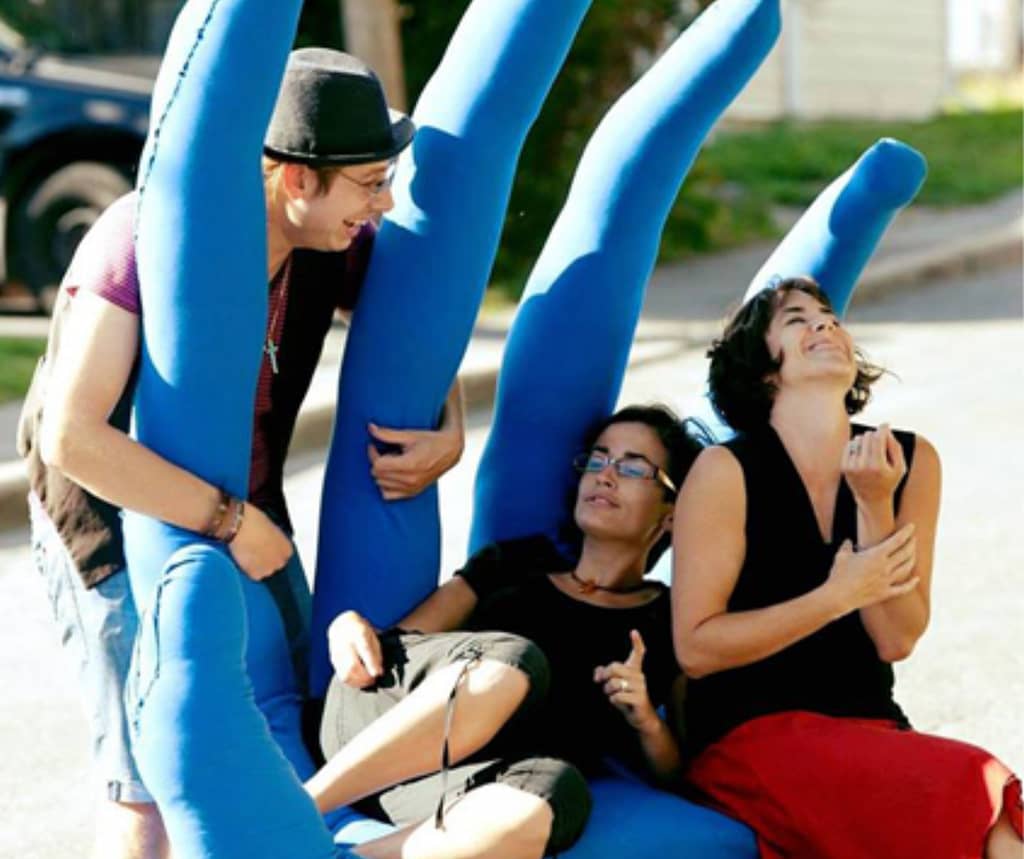 Description de l’image : Trois personnes s’appuient sur une géante main bleue en extérieur. La personne de gauche debout regarde les deux autres personnes assises et semi-allongées.