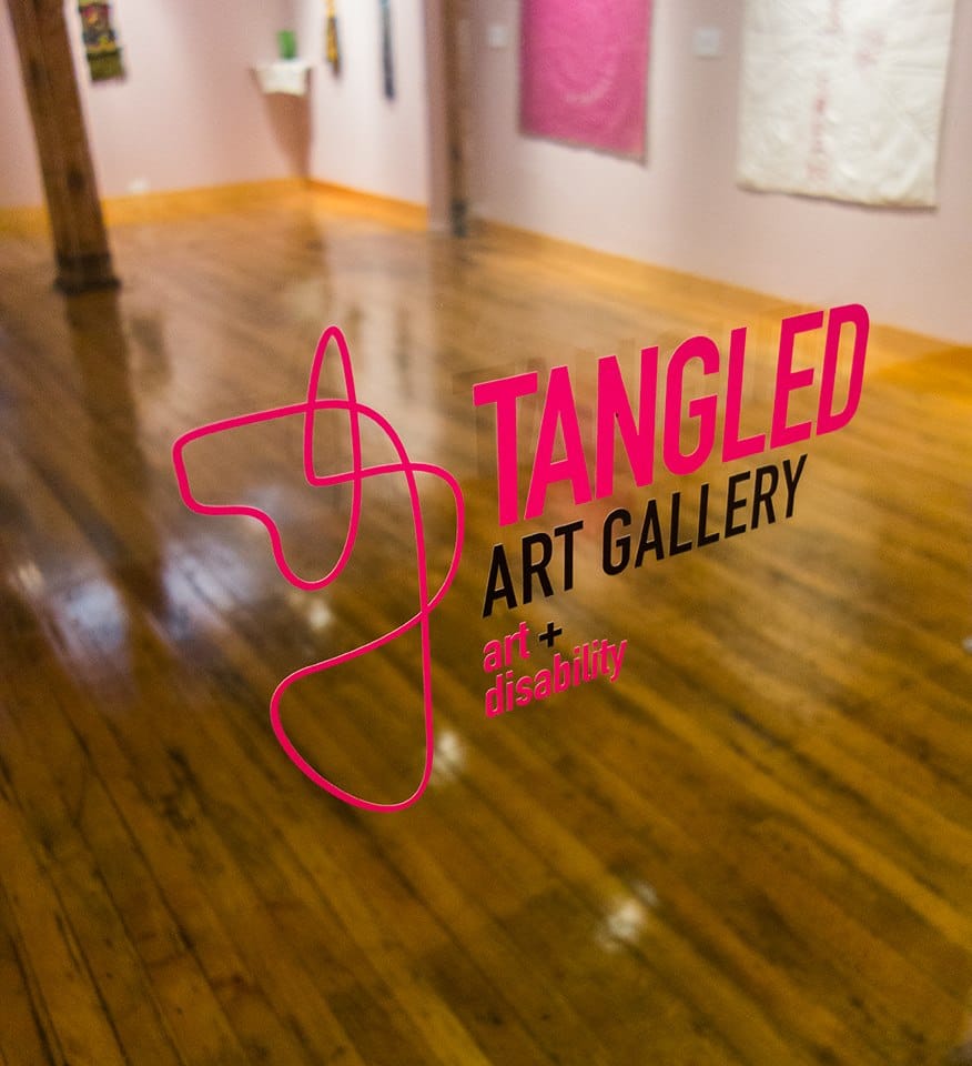 Description de l’image : sur une vitre transparente, il y a le logo rose et noir de la galerie Tangled Arts+ Gallery. En arrière-plan, c’est une salle avec du bois au sol et des œuvres accrochées au mur. 